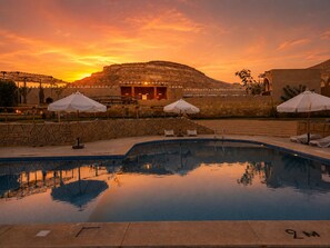 Zona de piscina con sillones de descanso y sombrillas, una pared de piedra y un edificio de techo plano al fondo.