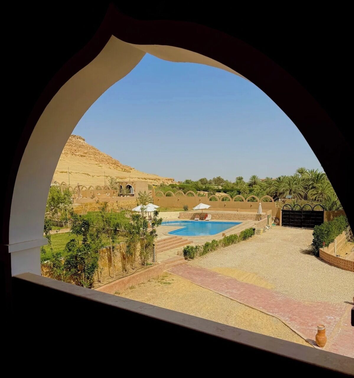Vista attraverso una finestra ad arco su un'area piscina con paesaggio desertico, palme e un edificio con archi.