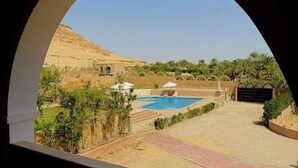 A view through an arched window of a pool area with a desert landscape, palm trees, and a building with arches. - Happy Oasis Hotel (Siwa)