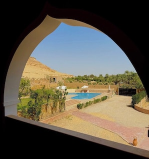 A view through an arched window of a pool area with a desert landscape, palm trees, and a building with arches. - Happy Oasis Hotel (Siwa)