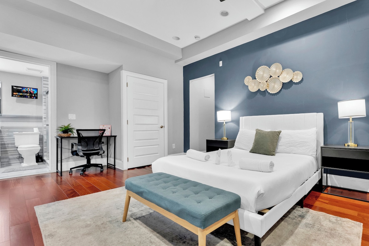 Un dormitorio moderno con una cama grande, dos mesitas de noche, un escritorio con silla y un baño visible a través de una puerta abierta.
