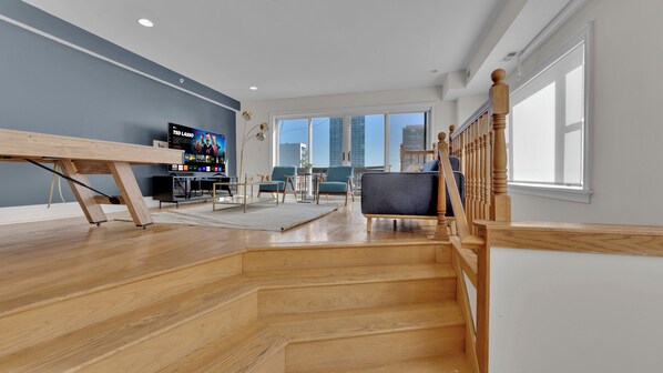 A spacious living area with a wooden staircase, a flat-screen TV, and a view of the city.