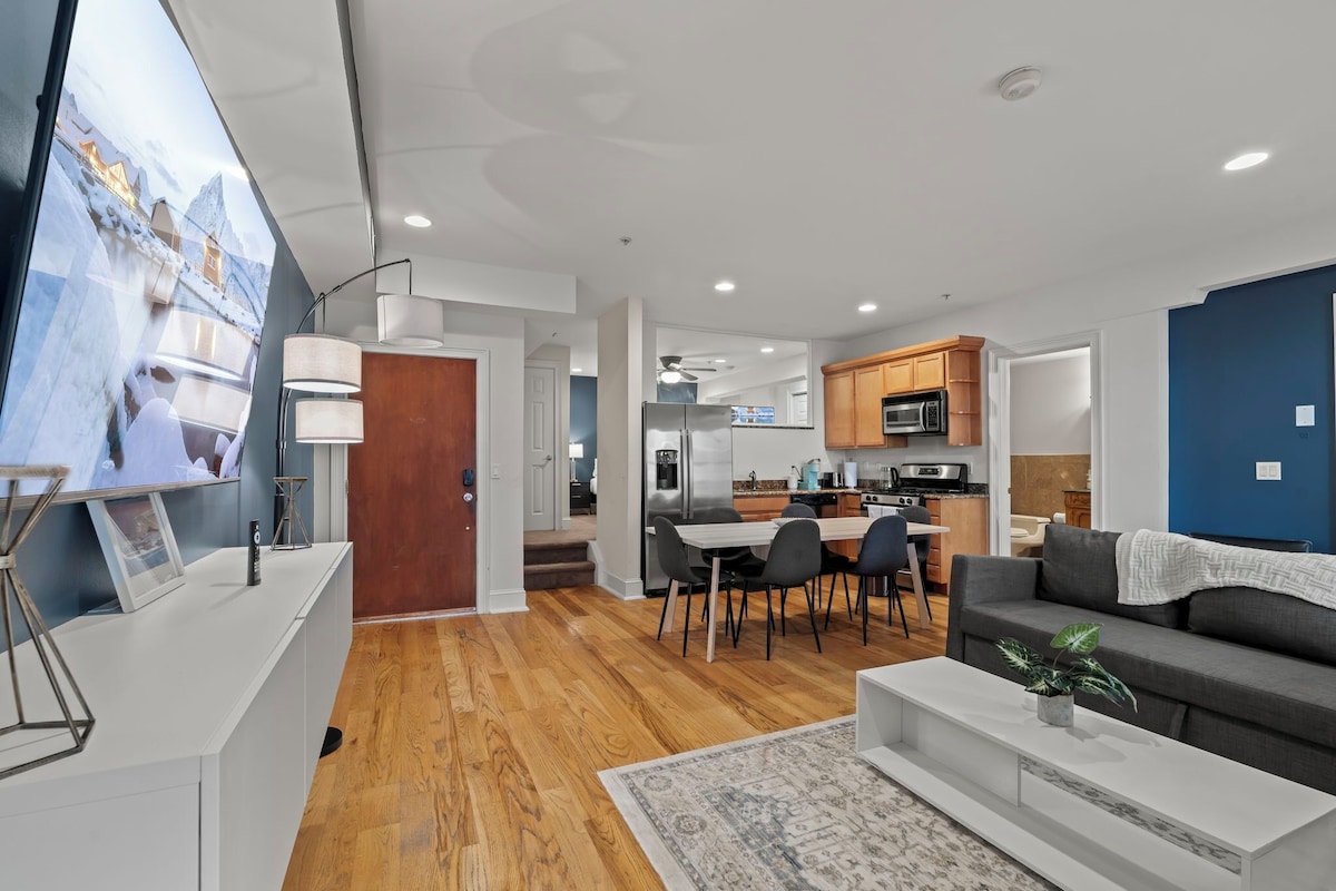 Una sala moderna con sofá, mesa de centro y televisor. Hay un comedor con sillas y una mesa, y una cocina con armarios y electrodomésticos.