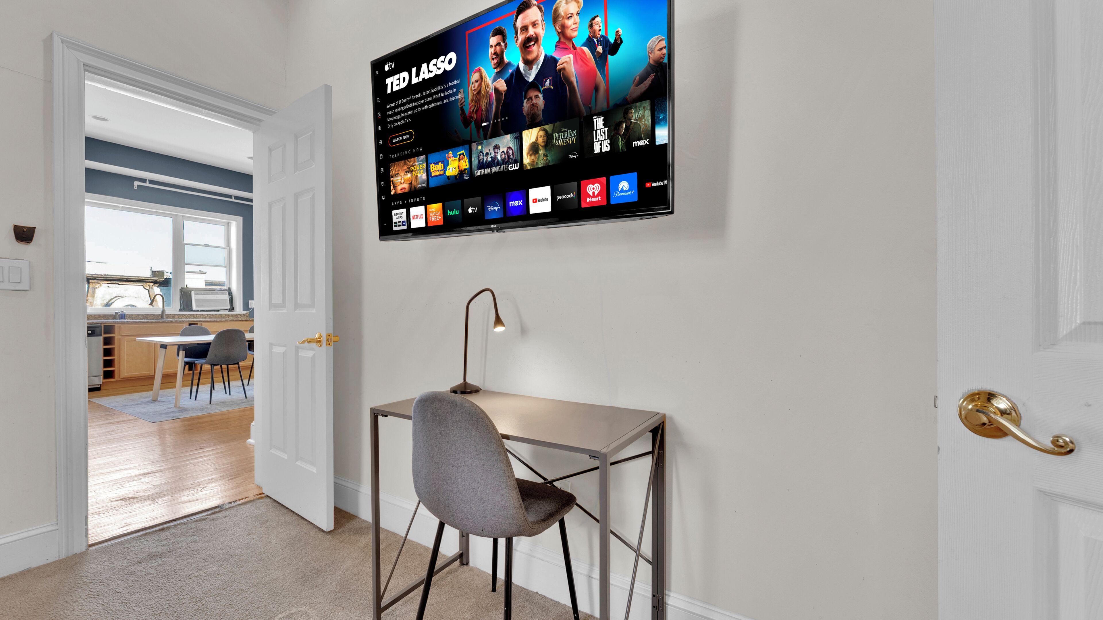 A modern living room with a flat-screen TV displaying a streaming service interface, a desk with a chair, and a door leading to a kitchen.