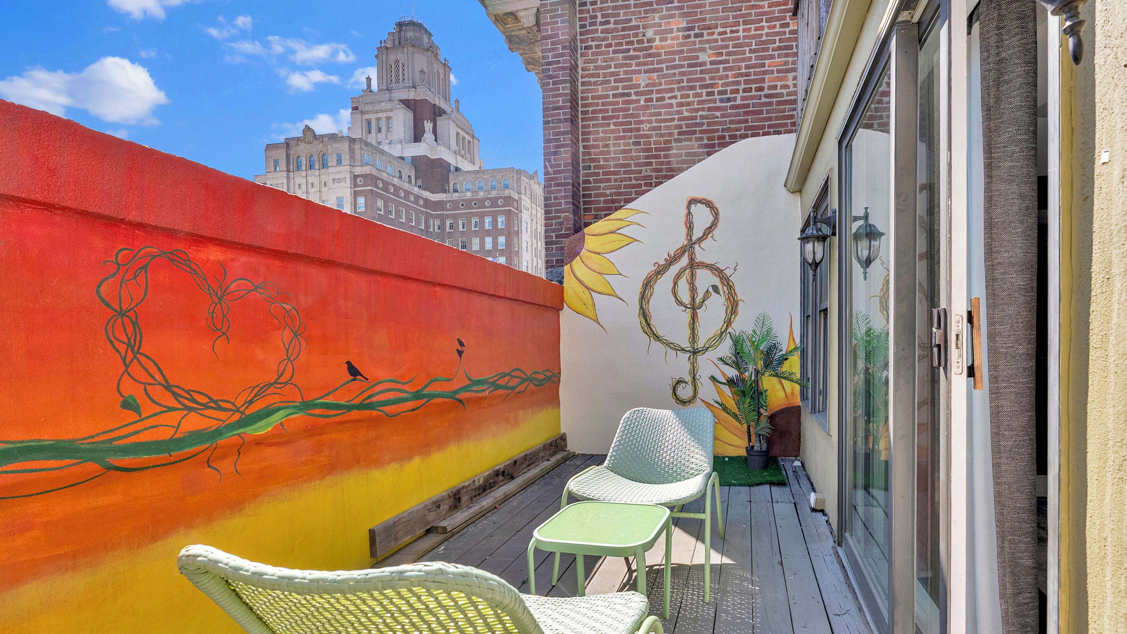A balcony with a painted wall, two wicker chairs, a small table, and a potted plant.