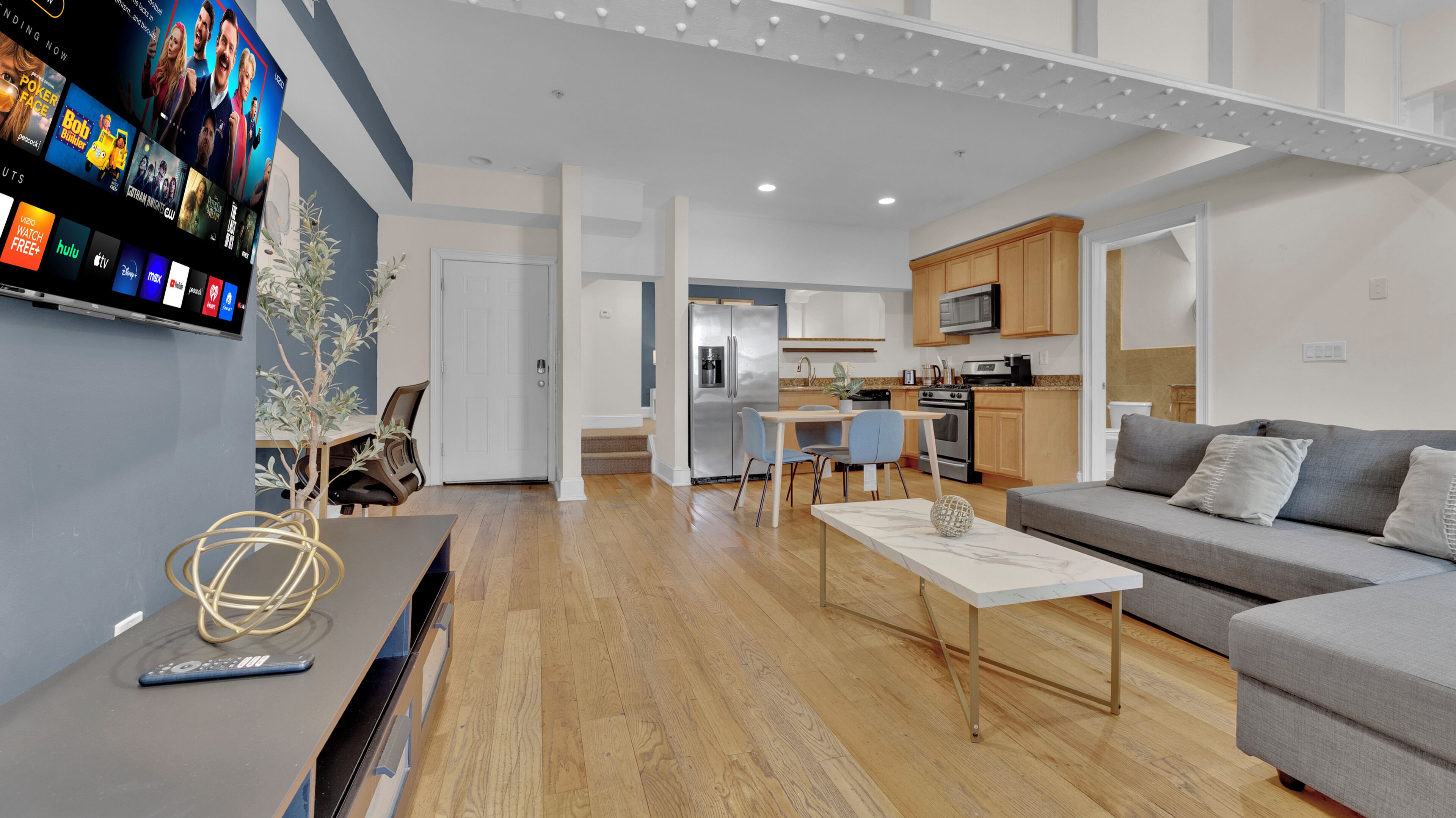 Un salon moderne avec un téléviseur à écran plat, un parquet en bois, un canapé gris et une cuisine en arrière-plan.