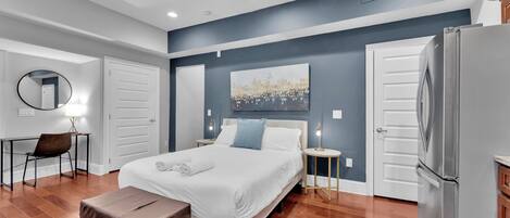 A bedroom with a bed, a desk with a chair, a round mirror, a bedside table with a lamp, and a refrigerator.