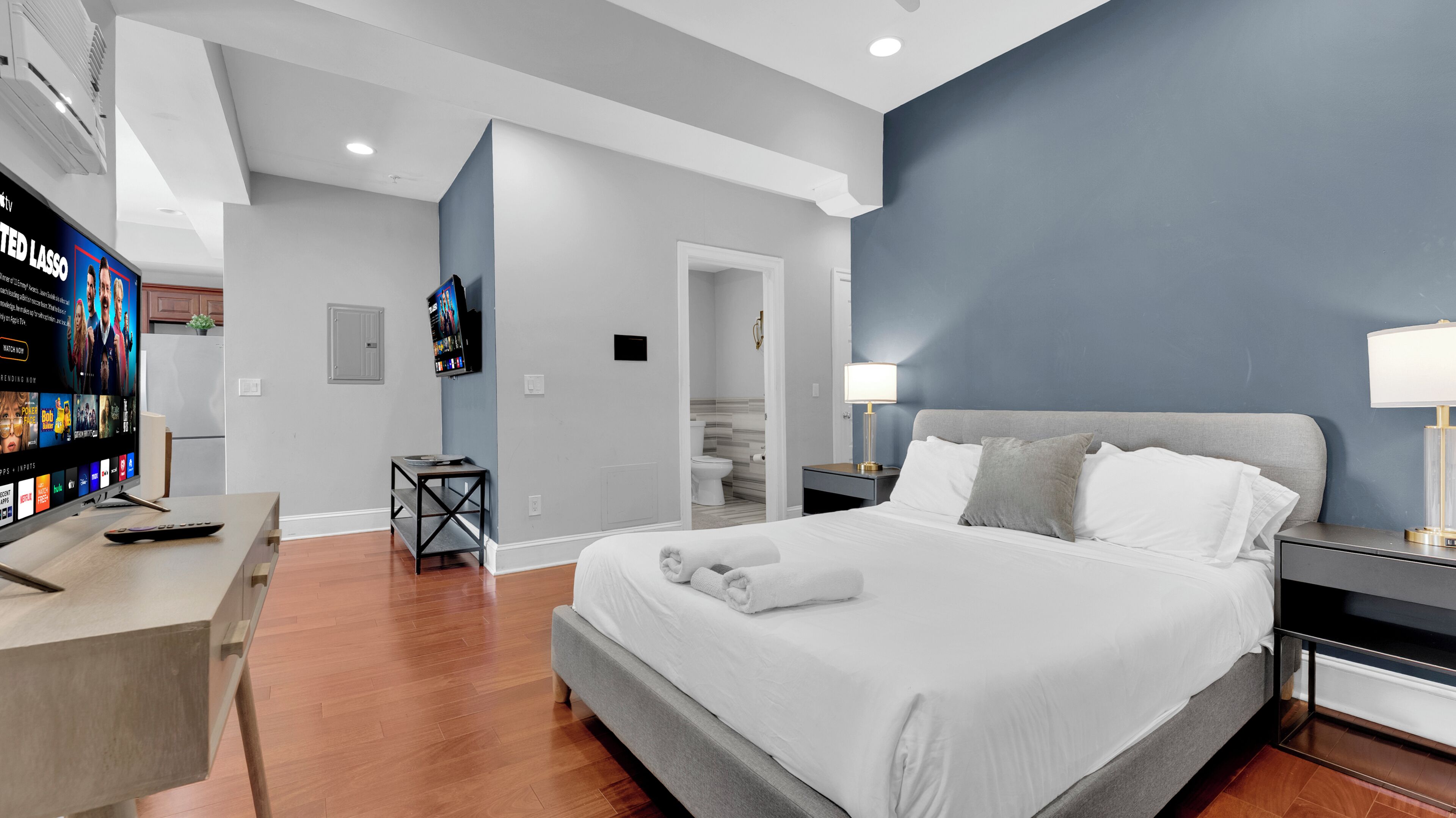 Quarto de hotel moderno com uma cama grande, mesas de cabeceira, uma escrivaninha com televisão e um banheiro visível por uma porta aberta.