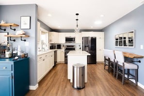 A modern kitchen with white cabinets, stainless steel appliances, a central island, and a dining area with chairs. - Sound View Retreat Near Illahee Beach & Ferry (Bremerton)