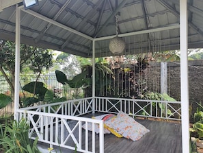 A covered outdoor seating area with a white railing, a hanging light fixture, and lush greenery in the background. - Cozzy Villa Swasti Cisarua (bogor)