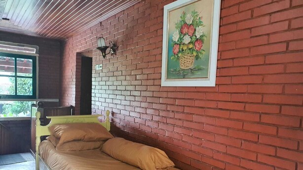 A single bed with a wooden frame and beige bedding in a room with red brick walls, a window, and a framed picture of flowers. - Cozzy Villa Swasti Cisarua (bogor)