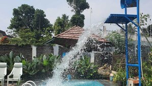 A swimming pool with a water fountain feature, surrounded by greenery and a tiled roof structure. - Cozzy Villa Swasti Cisarua (bogor)