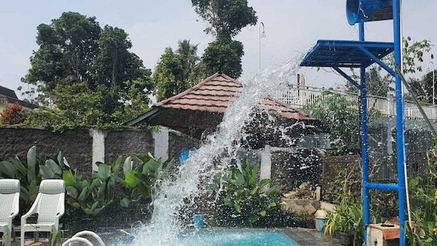 A swimming pool with a water fountain feature, surrounded by greenery and a tiled roof structure. - Cozzy Villa Swasti Cisarua (bogor)