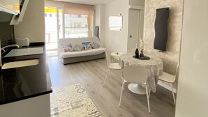 A modern kitchen with a black countertop, a dining area with a white table and chairs, a sofa with cushions, and a flat-screen TV mounted on the wall. - Sitges Oasis Apartment with Balcony (Sitges)