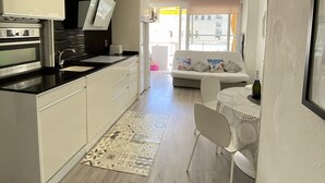 A modern kitchen with white cabinets, a black countertop, and a built-in oven. There is a dining area with a white table and chairs, and a sofa in the background. - Sitges Oasis Apartment with Balcony (Sitges)