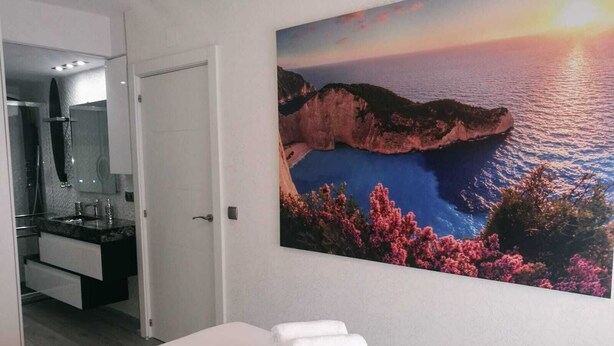 A hotel room with a bed, a bathroom with a sink and mirror, and a large scenic painting of a coastal landscape. - Sitges Oasis Apartment with Balcony (Sitges)