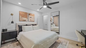 Ein modernes Schlafzimmer mit Bett, Nachttisch, Lampe, Deckenventilator, Kunstwerk, Spiegel und Schreibtisch mit Stuhl.