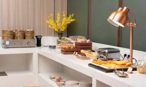 Tarjolla on monipuolinen buffet, jossa on erilaisia ruokia, kahvinkeitin ja kuparinen lamppu.