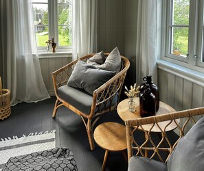 Ein Korbstuhl mit Kissen, ein kleiner runder Tisch mit einer Flasche und Blumen, und Blick auf Grün hinter einem Fenster.