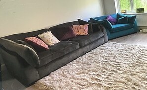 Ein dunkelbraunes Sofa mit gemusterten Kissen, ein heller HochflorTeppich und ein Türkisblaues Sofa ebenfalls mit Kissen in einem Raum mit einem Fenster.