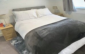Ein Schlafzimmer mit Bett, Nachttisch, Lampe, Kommode und einem Fenster mit Vorhängen.