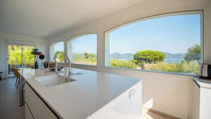 Cocina moderna con una isla grande, armarios blancos y amplios ventanales con vistas a zonas verdes y colinas.
