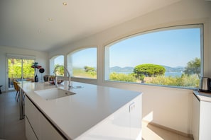 A modern kitchen with a large island, white cabinetry, and large windows offering a view of greenery and hills.