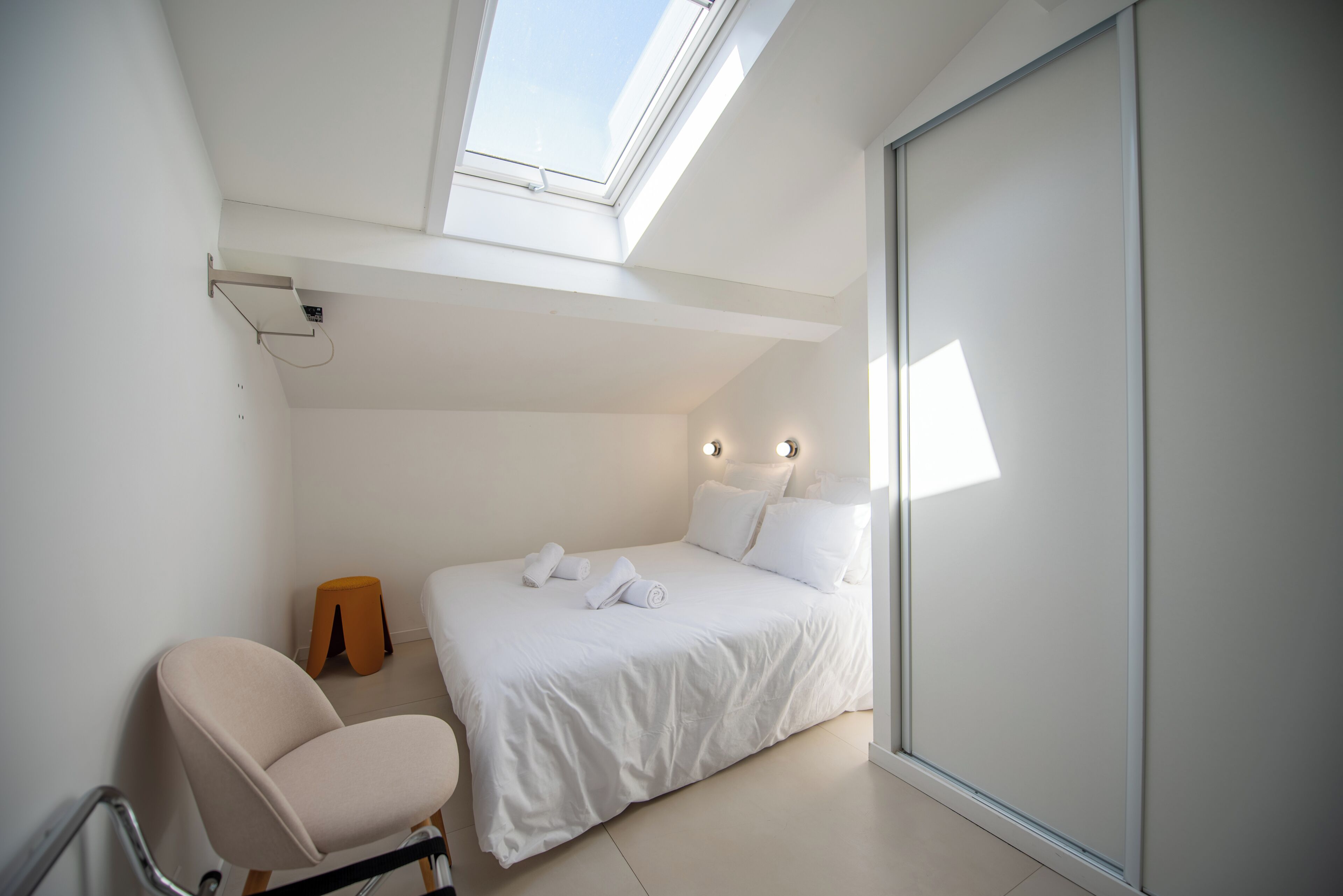 A bedroom with a skylight, a bed with white linens, a chair, and a small stool.