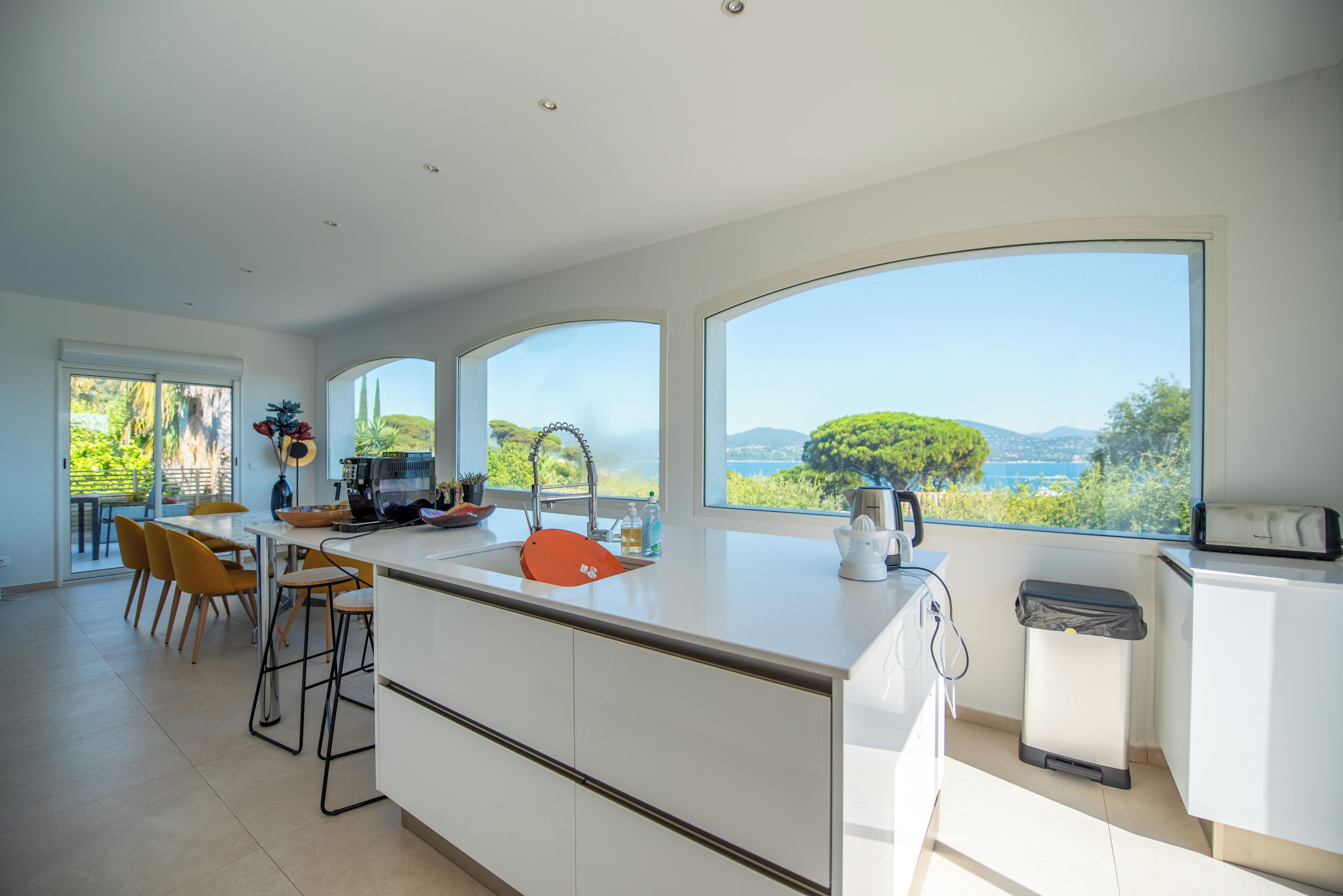 Una cocina moderna con una isla grande, un comedor con sillas amarillas y amplios ventanales con vistas a zonas verdes y colinas.