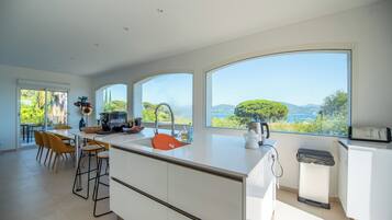 Una cocina moderna con una isla grande, un comedor con sillas amarillas y amplios ventanales con vistas a zonas verdes y colinas.