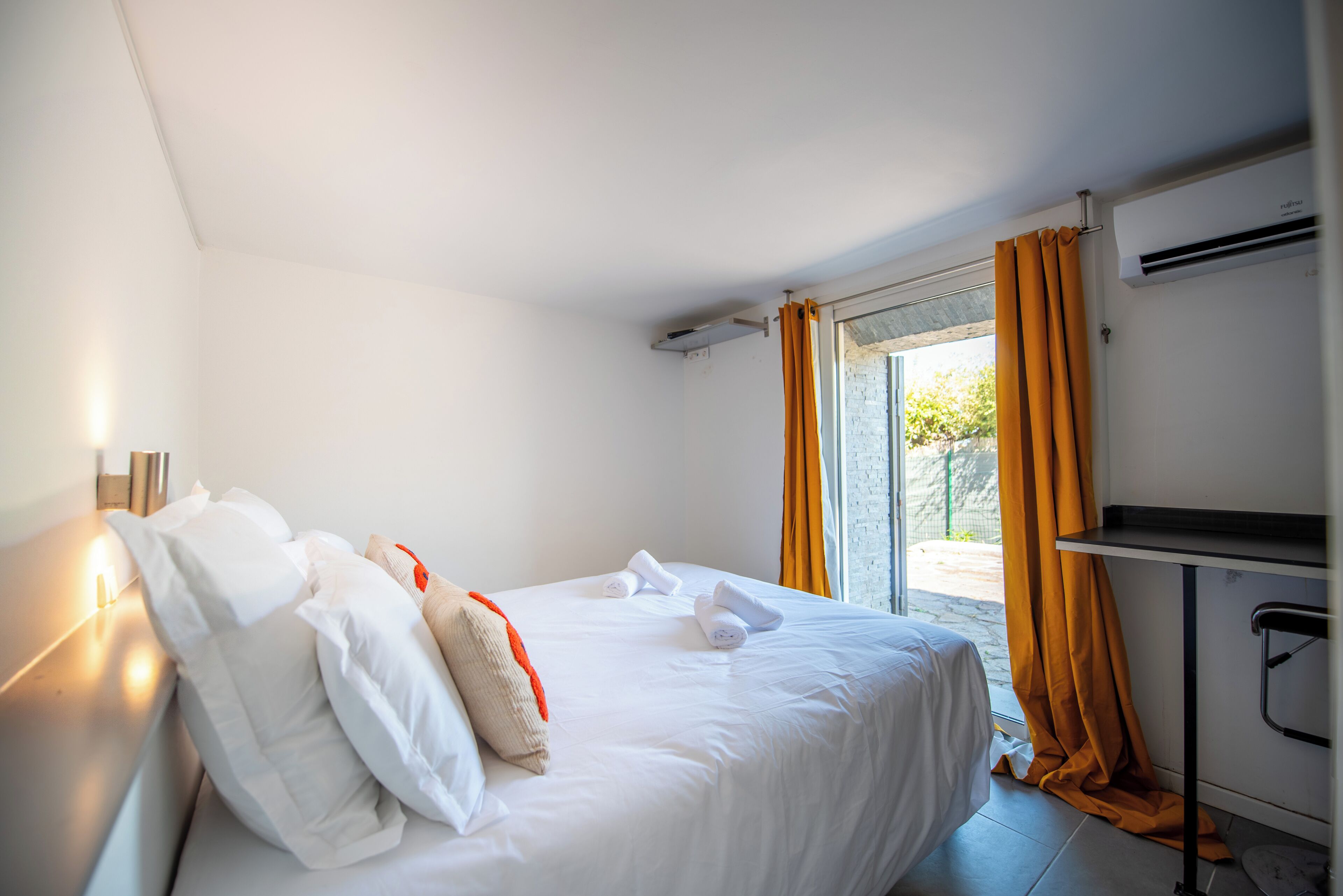 A hotel room with a bed, a desk, an air conditioning unit, and a view through the window.