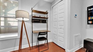 Una habitación con una lámpara de pie, una estantería metálica, una silla, una puerta blanca y una pantalla montada en la pared.