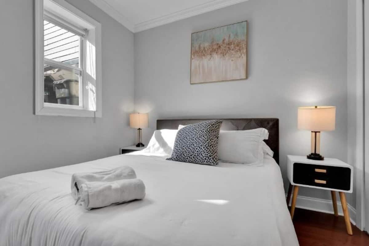 A bedroom with a large bed, two bedside tables, a window, and a painting on the wall.
