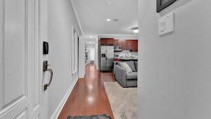 Un pasillo que lleva a una cocina con piso de madera, un sofá gris y un refrigerador.