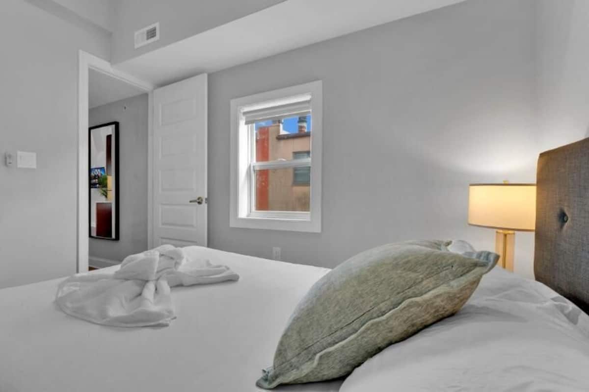 A hotel room with a bed, a window, a lamp, and a door.