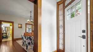 A wooden-floored hallway leading to a white door with a stained glass window, a dining area with floral upholstery, and a chandelier. - Charming & Updated 1867 Brick House in Historic Downtown Delaware, OH; Sleeps 12 (Delaware)