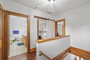 A hallway with wooden flooring, a white wall, a wooden railing, and a door leading to a bathroom.