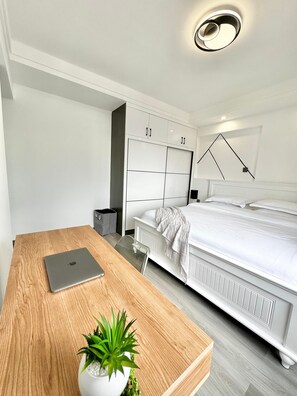 Een moderne slaapkamer met een houten bureau, een laptop, een potplant, een bed met witte beddengoed en een witte kast.