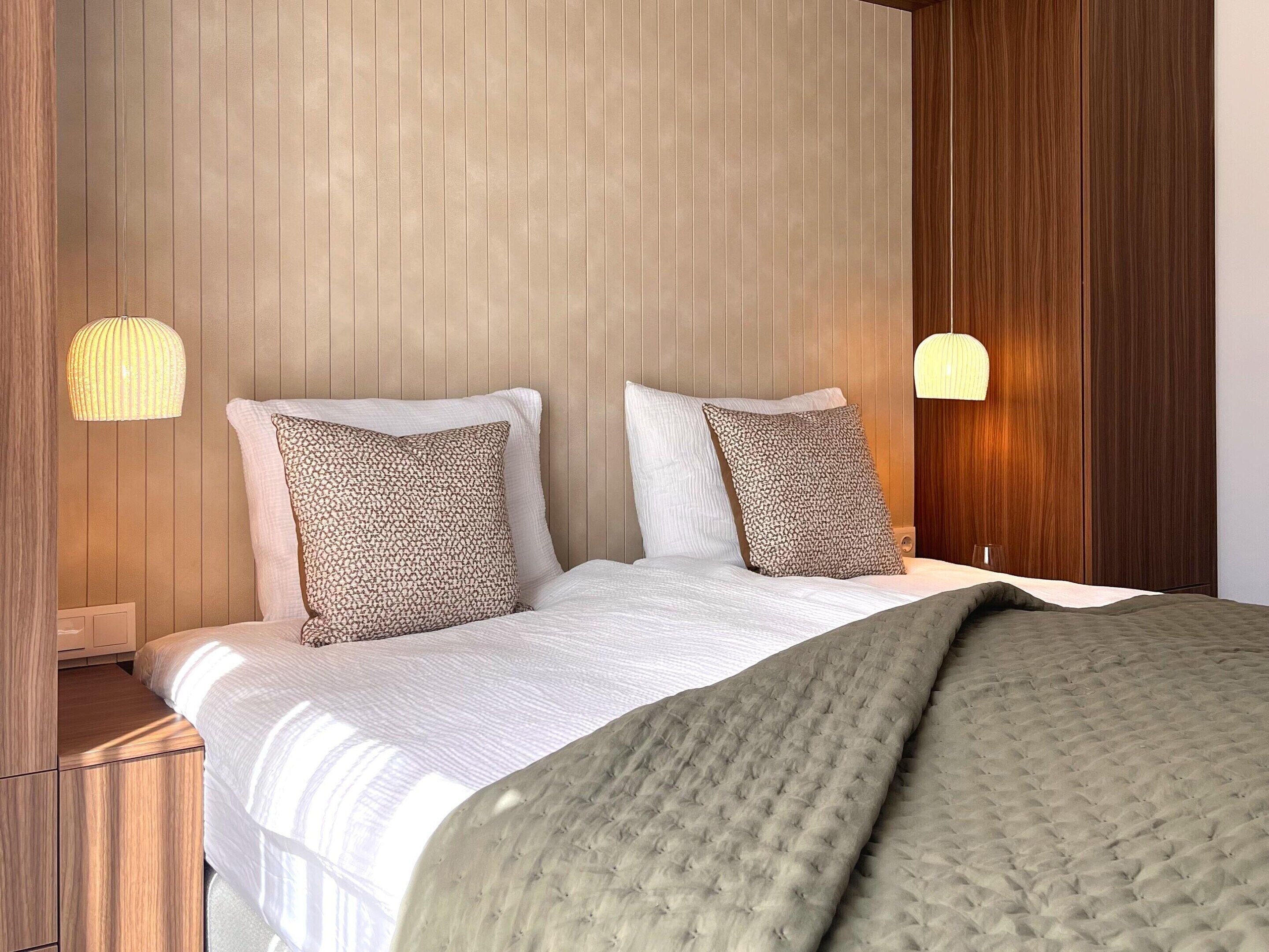 A bed with a white duvet, two patterned pillows, a green throw blanket, and wooden paneling on the walls.