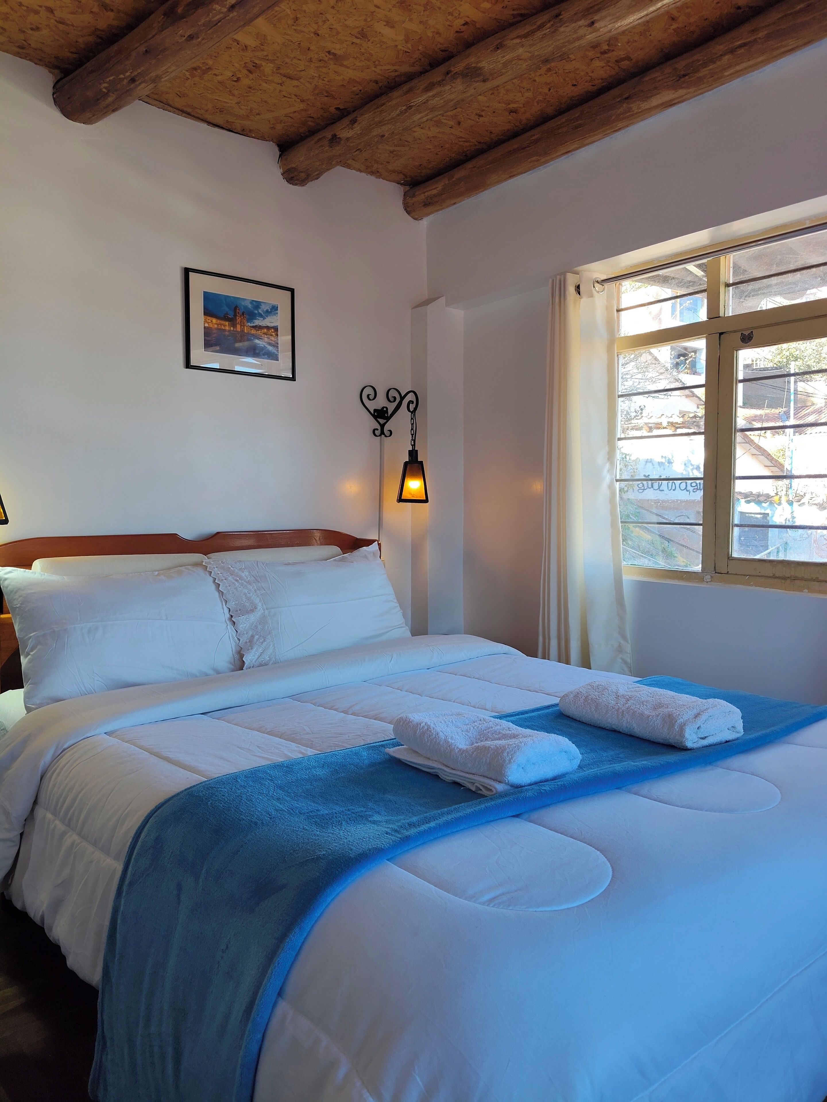 Ein ordentlich gemachtes Bett mit weißen Laken, einer blauen Decke und zwei gefalteten Handtüchern. Über dem Bett hängt ein gerahmtes Bild.