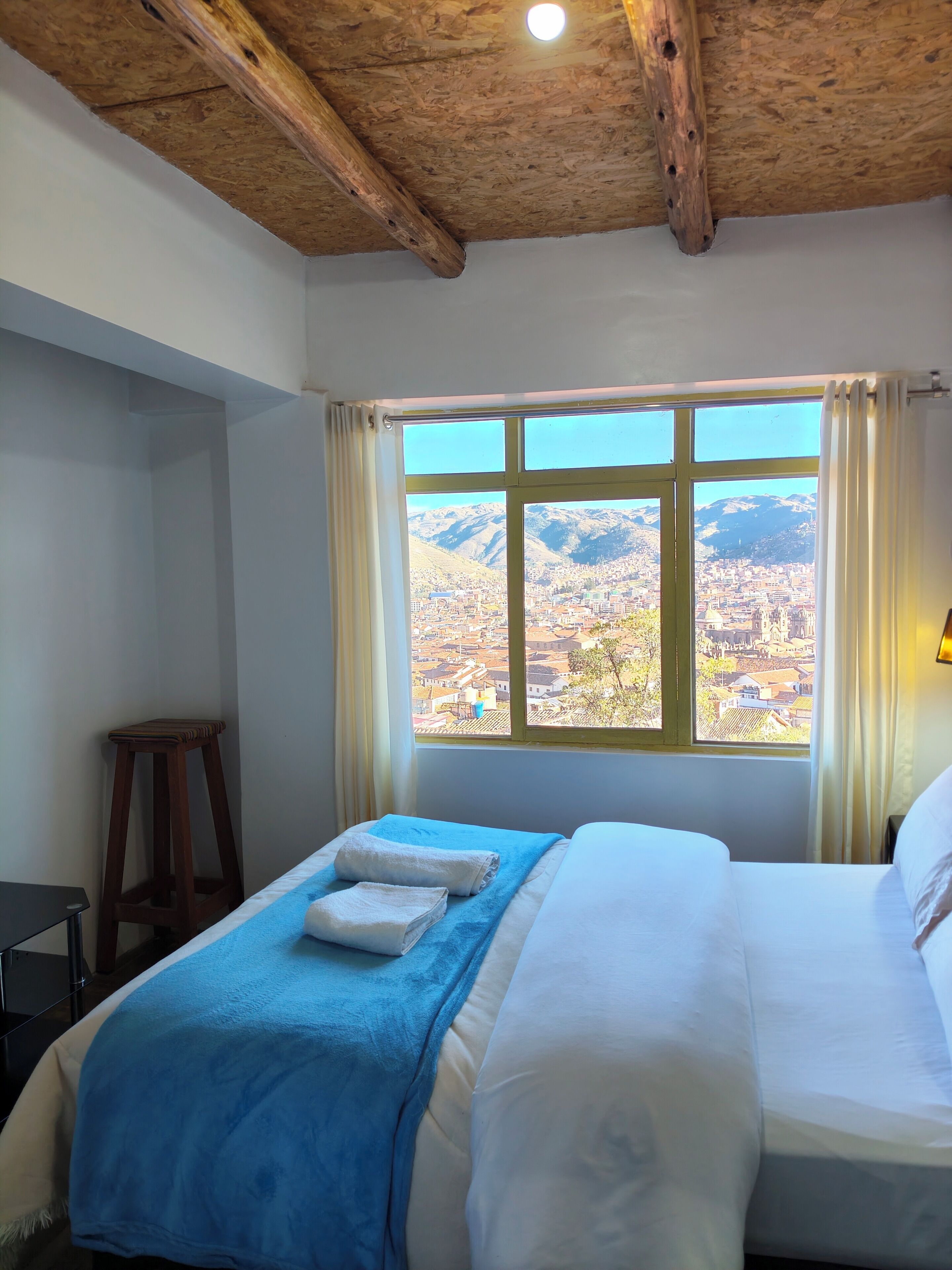Une chambre d’hôtel avec une grande fenêtre, des poutres en bois au plafond, un lit avec du linge de lit blanc et une couverture bleue.