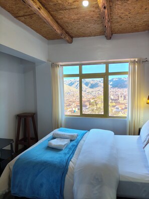A hotel room with a large window, wooden ceiling beams, a bed with white linens, and a blue blanket. - Serene 2-bedroom apartment in charming Cusco perfect for explorers (Cusco)