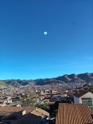 Eine Stadtansicht mit blauem Himmel, einem deutlichen Mond und einem Bergpanorama im Hintergrund.