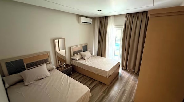 A hotel room with two beds, a bedside table, a mirror, an air conditioning unit, and a view of the outside through a window with curtains.