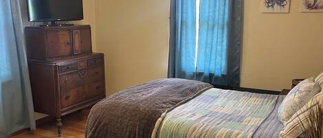 Un dormitorio con cama, cómoda, televisor, ventilador de techo y cortinas.