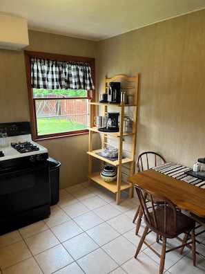 A kitchen with a stove, a wooden shelf with kitchenware, a dining table with chairs, and a window with a view of a backyard.