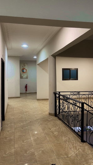 A corridor with a tiled floor, a fire extinguisher, a ceiling light, and a window with a metal railing. - Maya Lodge (Accra)