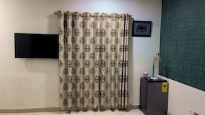 A room with a patterned curtain, a flat-screen TV mounted on the wall, a framed picture, and a desk with a lamp and a small pink object. - Maya Lodge (Accra)