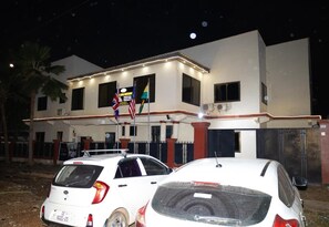 A two-story building with a gated entrance, a flag, and two parked cars. - Maya Lodge (Accra)