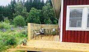 A wooden deck with outdoor furniture, a red building with a window, and a forested background.